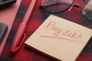 Close-up of a note reading 'Pay debt' next to a red pen on a plaid fabric, emphasizing financial reminders.